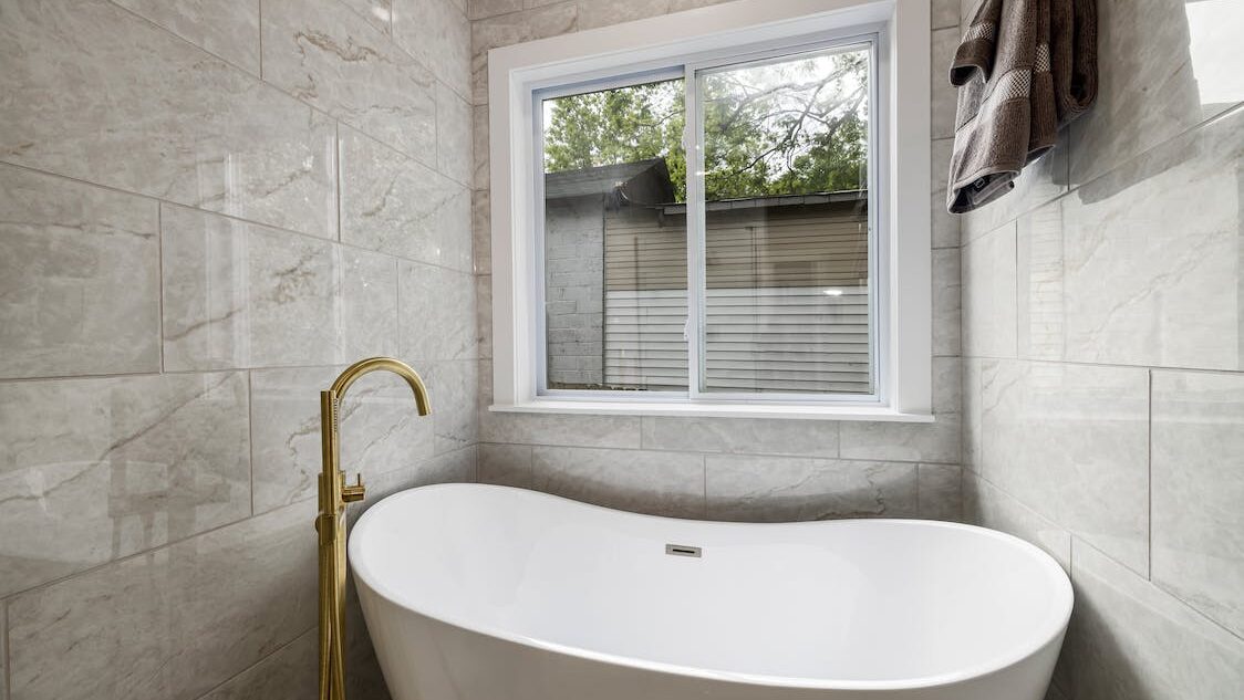 Freestanding bathtub in a bright tiled bathroom with large window and gold faucet